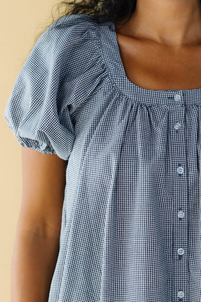 Close view of a woman wearing a navy & white gingham button down dress with a scoop neckline and large puff sleeves.