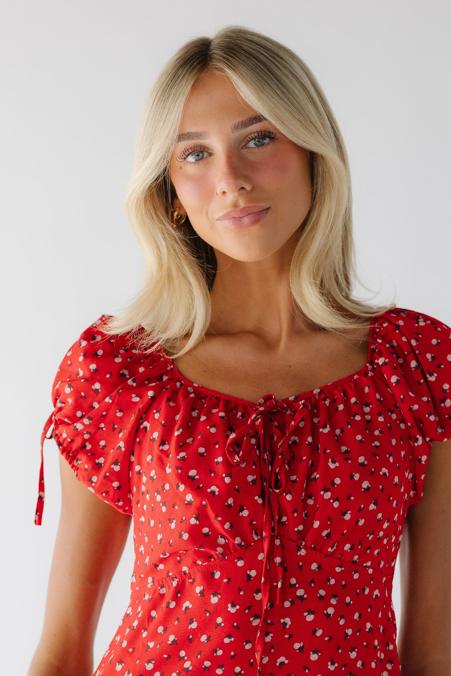 Close view of a woman wearing a red floral print dress with an empire waist, short puff sleeves and a drawstring scoop neckline.