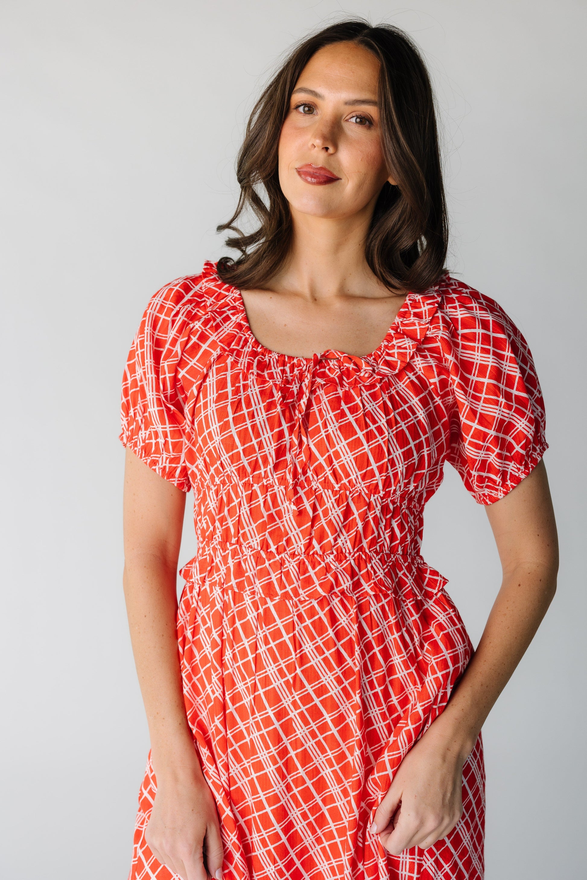 Close view of a woman wearing a red and white plaid dress with a scoop neckline, short puff sleeves and a wide accentuated  waist with a ruffle below the waistband.