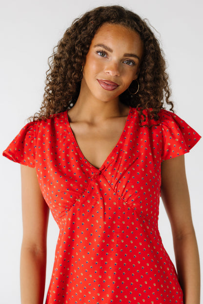 Close view of a teen girl wearing a red print dress with a pointed empire waist, V-neckline and short flutter sleeves.