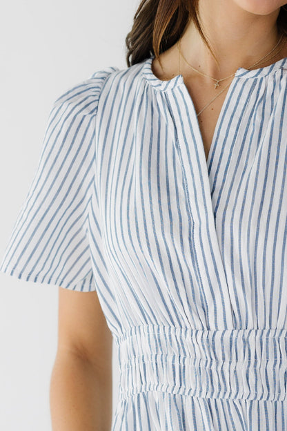 Close view of a young woman wearing a white and blue stripe dress with a wide elastic waistband, V-neckline and short sleeves.