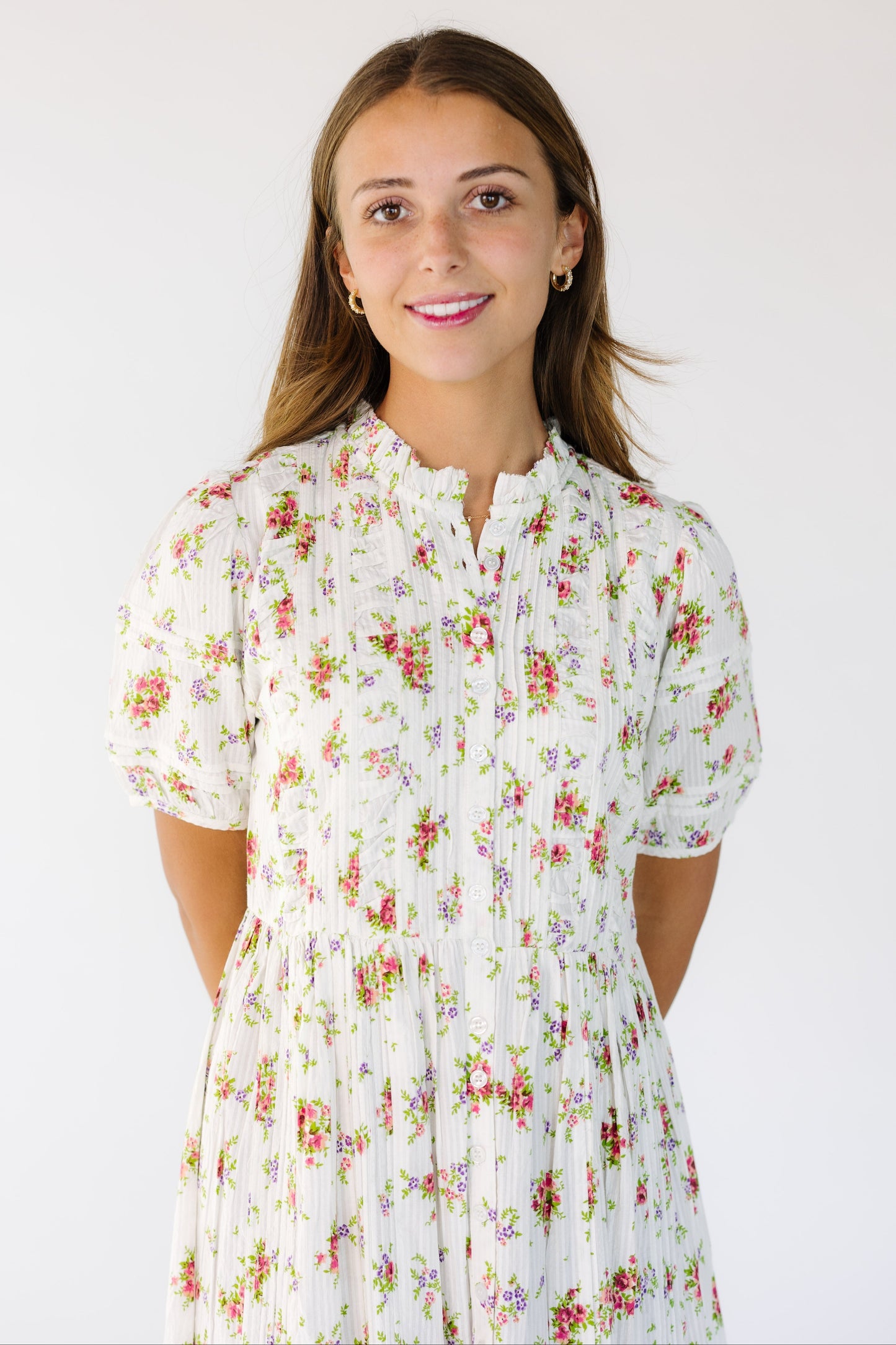 Close view of a young woman wearing a white and rose floral print dress with a high round collar with a stand up ruffle and buttons down the full length of the dress.
