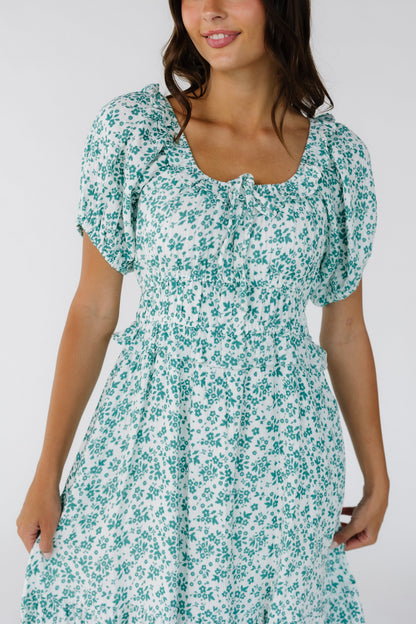 Close view of a woman wearing a white with teal floral print with a ruffled scoop neckline, short puff sleeves and a wide waistband.