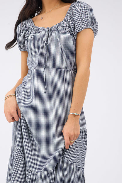 Close up view of a woman wearig a navy  white gingham midi dress with short puff sleeves and a drawstring scoop neckline.