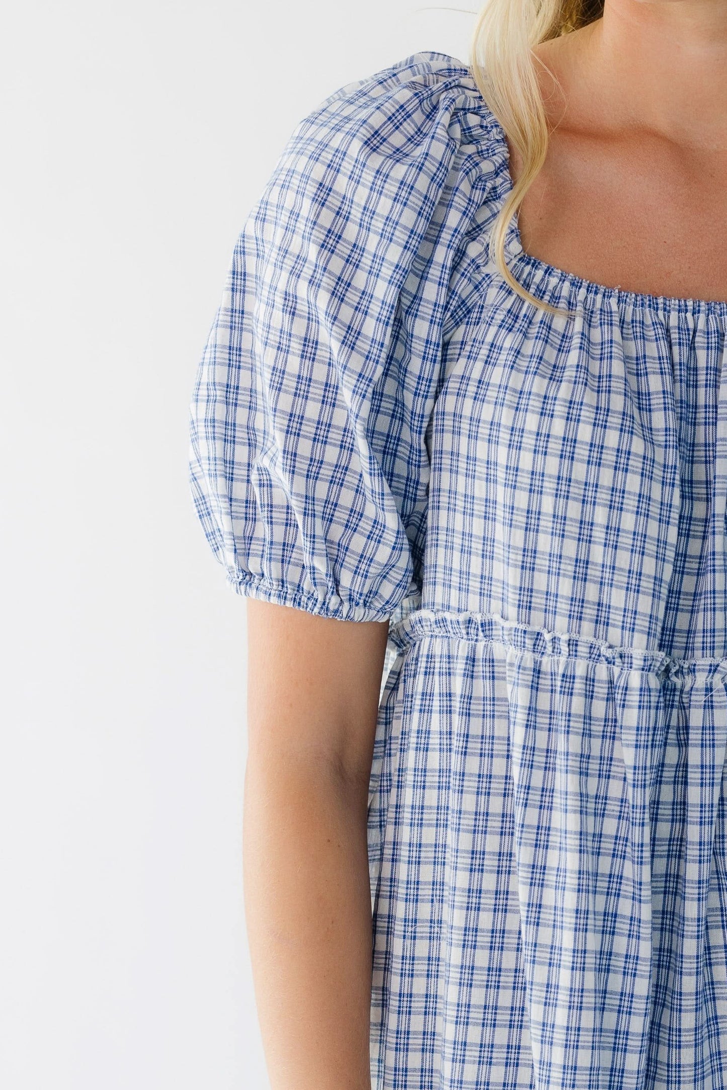 Close view of a woman wearing a blue plaid dress with a square neckline, large puff sleeves and tieres.