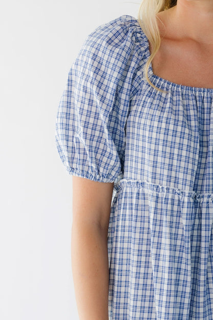 Close view of a woman wearing a blue plaid dress with a square neckline, large puff sleeves and tieres.