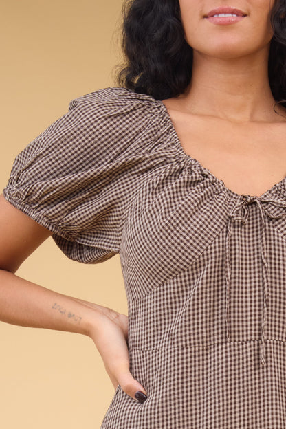 Close view of a woman wearing a brown gingham dress wtih short puff sleeves and a drawstring scoop neckline.