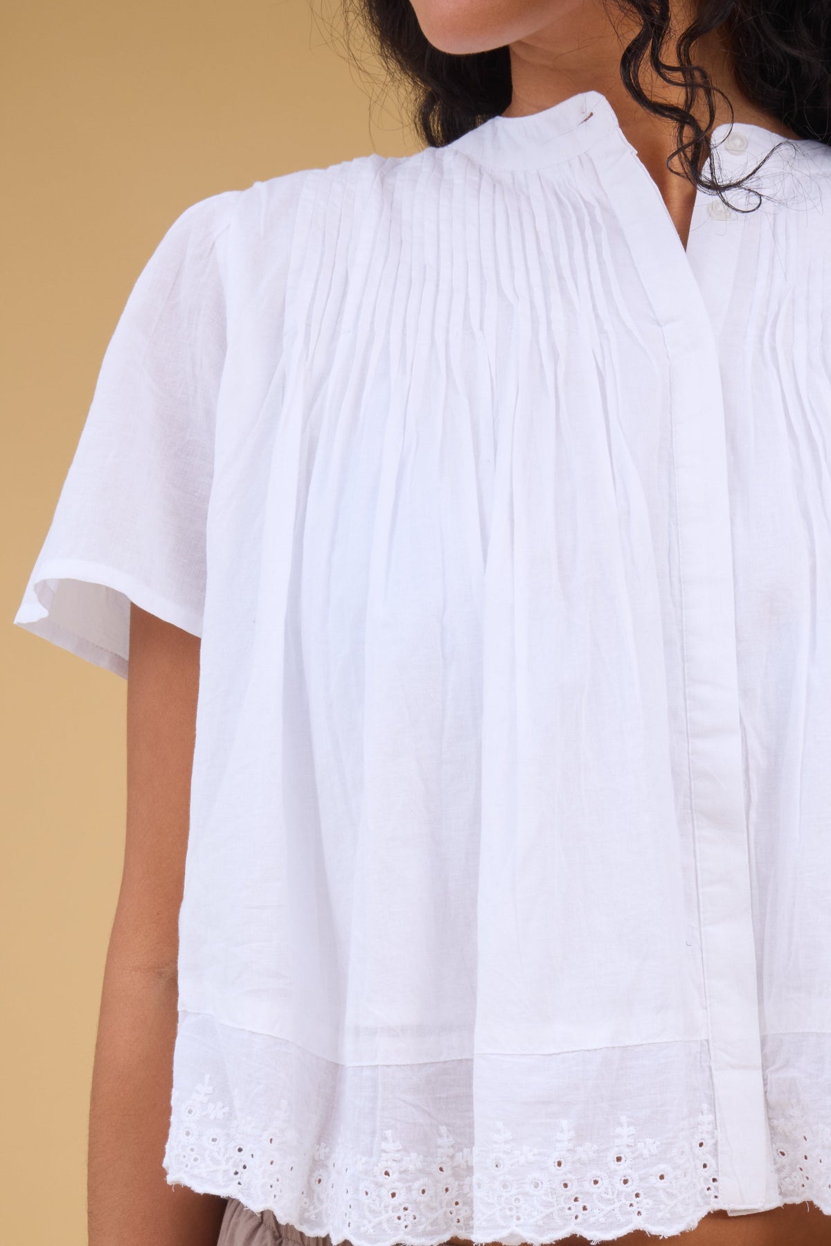 Close view of a woman wearing a loose fitting button down blouse in white with a hidden button down placket, pintucks at the shoulders and short sleeves.