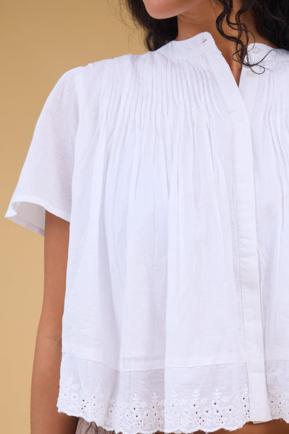 Close view of a woman wearing a loose fitting button down blouse in white with a hidden button down placket, pintucks at the shoulders and short sleeves.