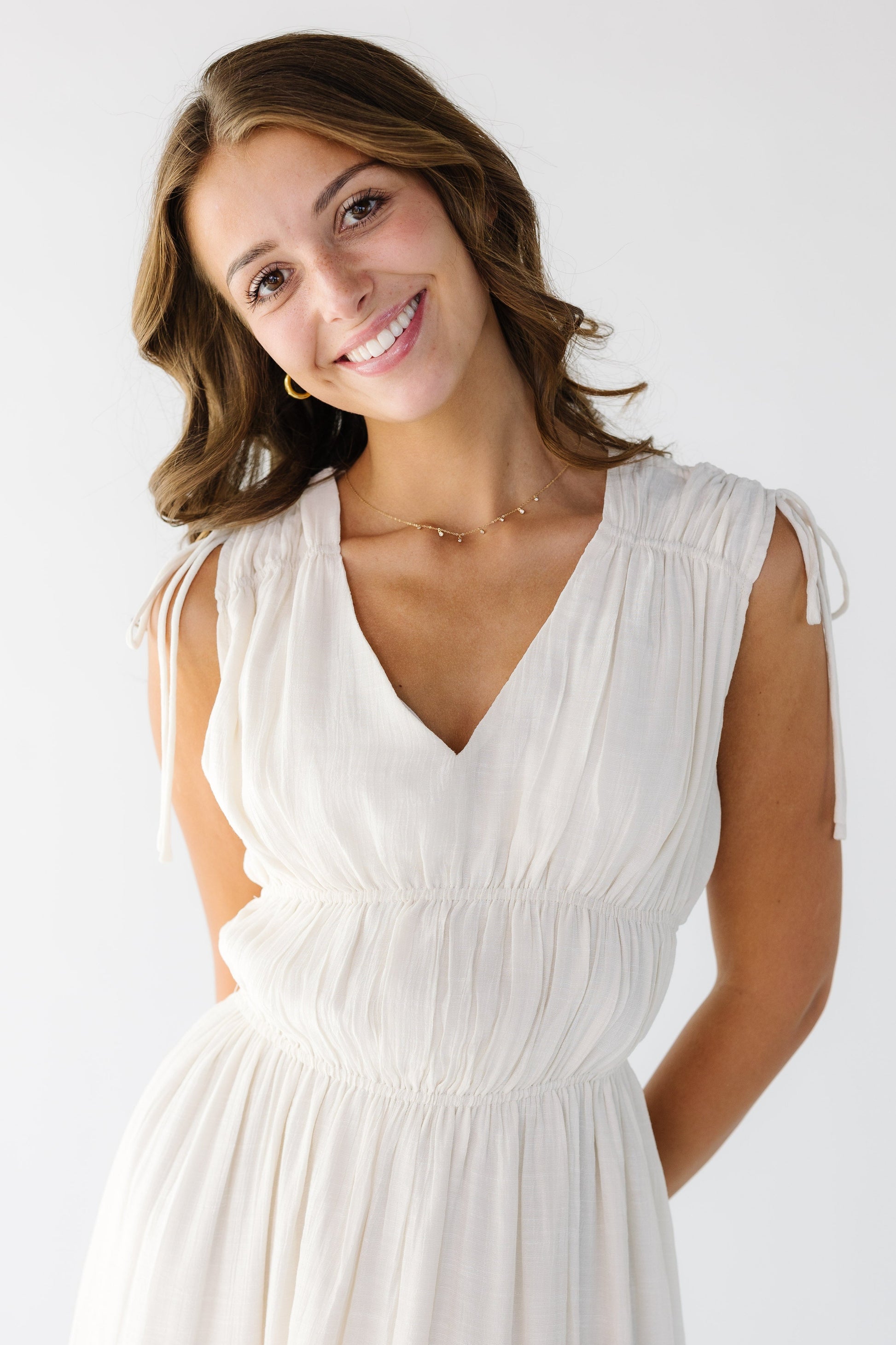 Close up view of a woman wearing a sleeveless  off white gauzy dress, with drawstring shoulders, V-neckline and bodice elastic gather details.