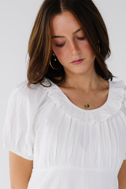 Close view of a woman wearing a white on white stripe dress with an empire waist, short puff sleeves and a scoop neckline with an eyelet ruffle.