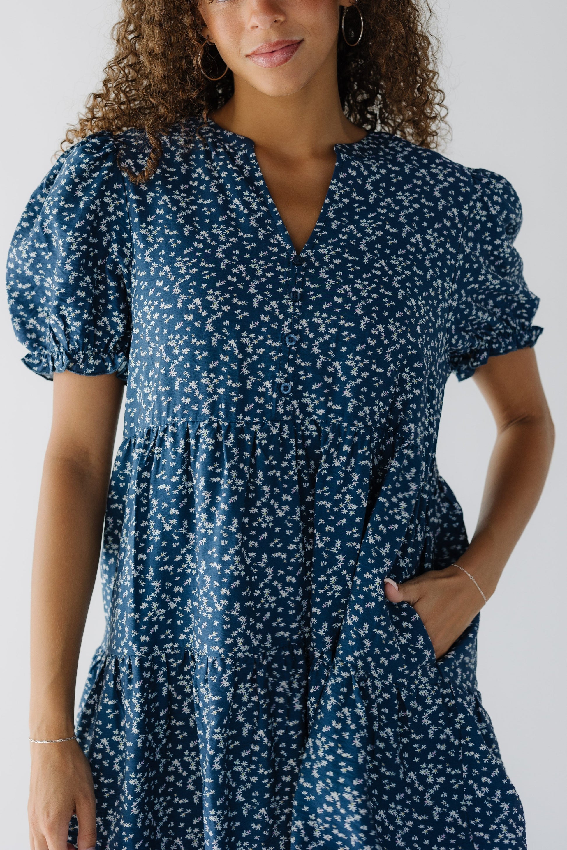 Close view of a teenage girl wearing a blue floral dress with side pockets, short puff sleeves and a V-neckline.