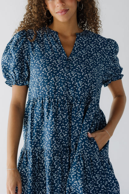 Close view of a teenage girl wearing a blue floral dress with side pockets, short puff sleeves and a V-neckline.