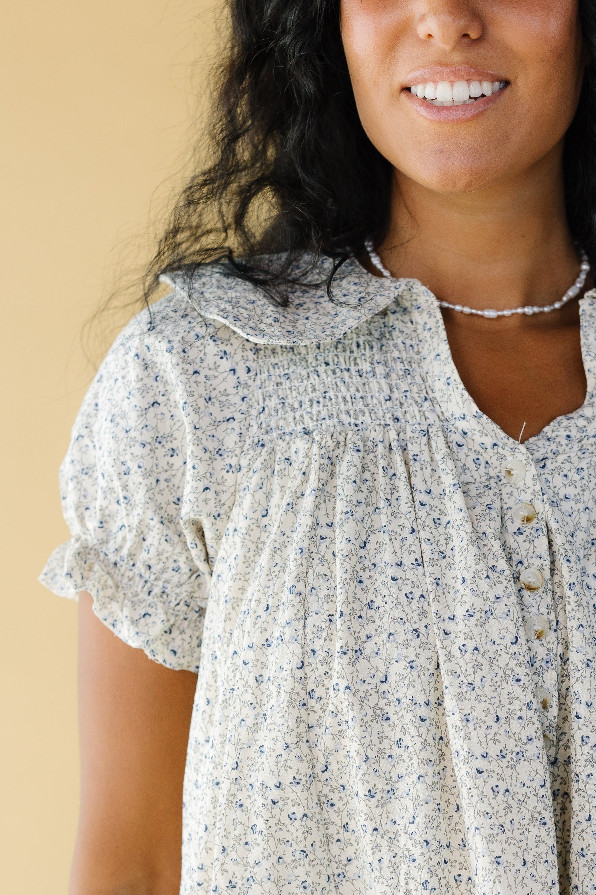 Close view o f a woman wearing a light grey and blue floral print dress with a short button placket, short sleeves, wide collar and smocked yoke.