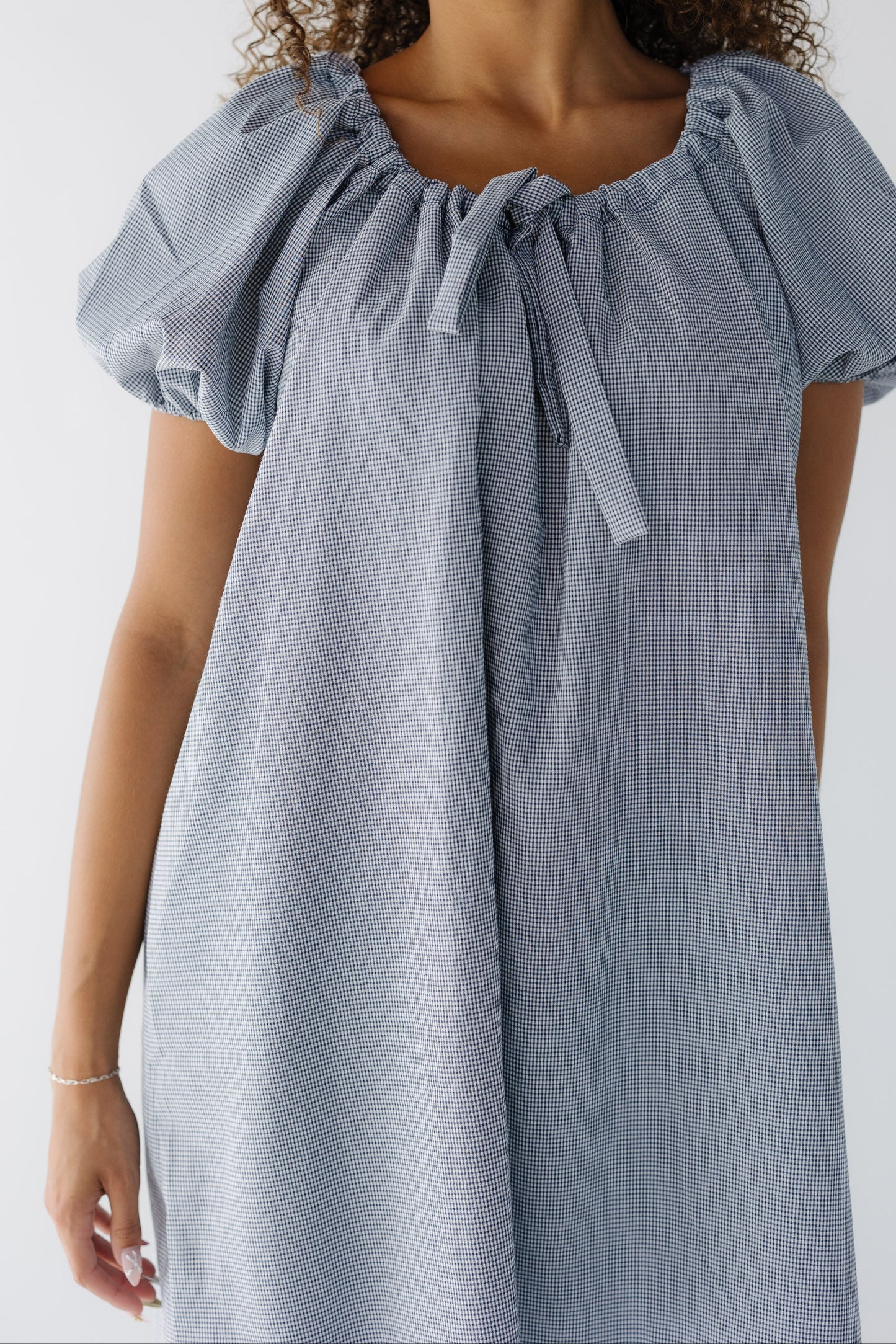 Closer view of a woman wearing a navy & white gingham short dress with balloon sleeves and a drawstring-scoop neckline.
