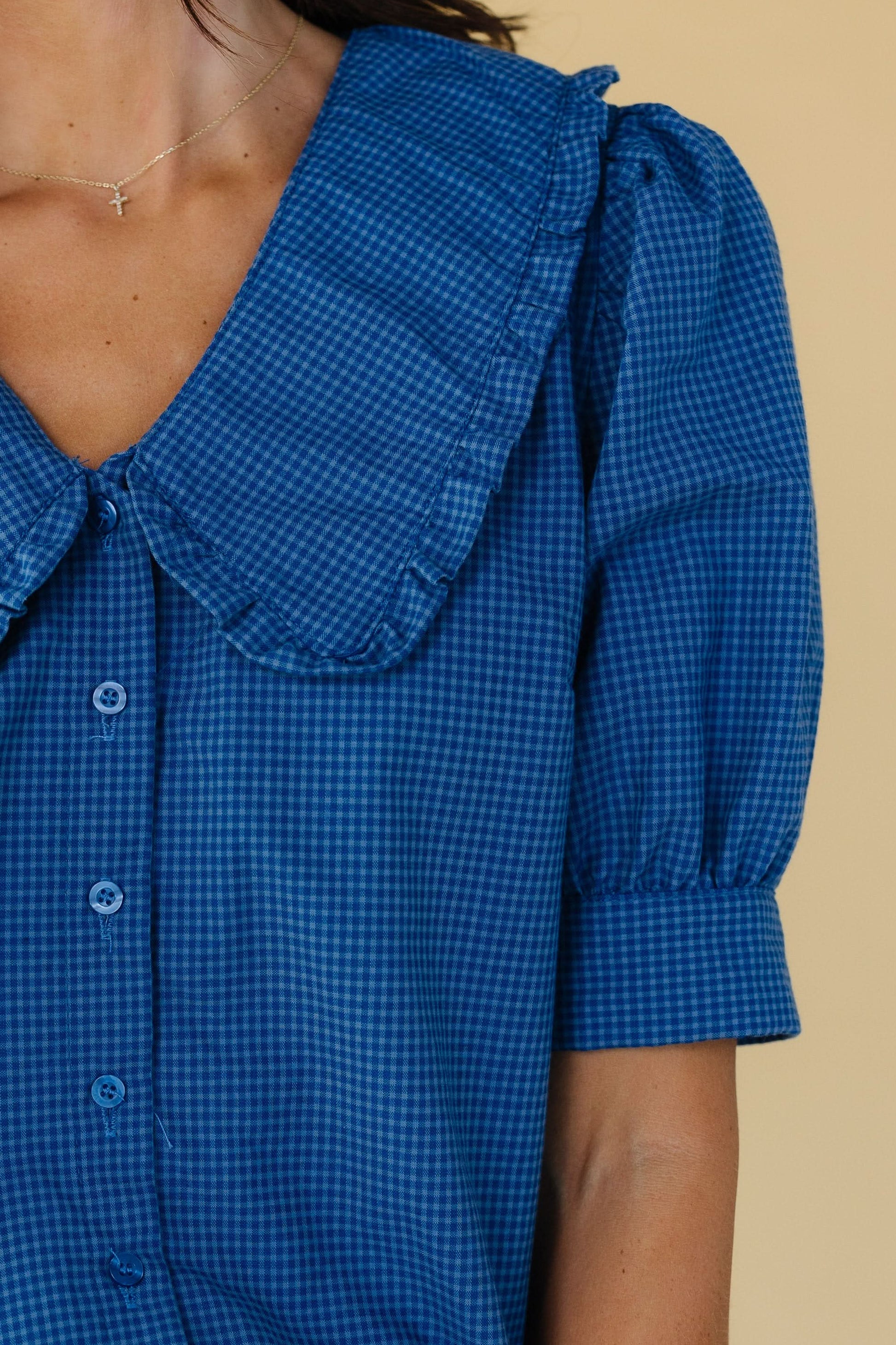 Close view of a woman wearing a blue gingham button down blouse with a V-neckline and wide ruffle edge collar and short puff sleeves with a wide cuff.