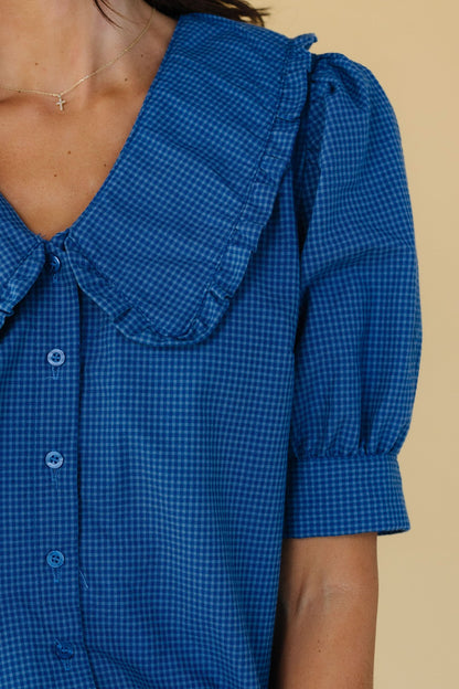 Close view of a woman wearing a blue gingham button down blouse with a V-neckline and wide ruffle edge collar and short puff sleeves with a wide cuff.