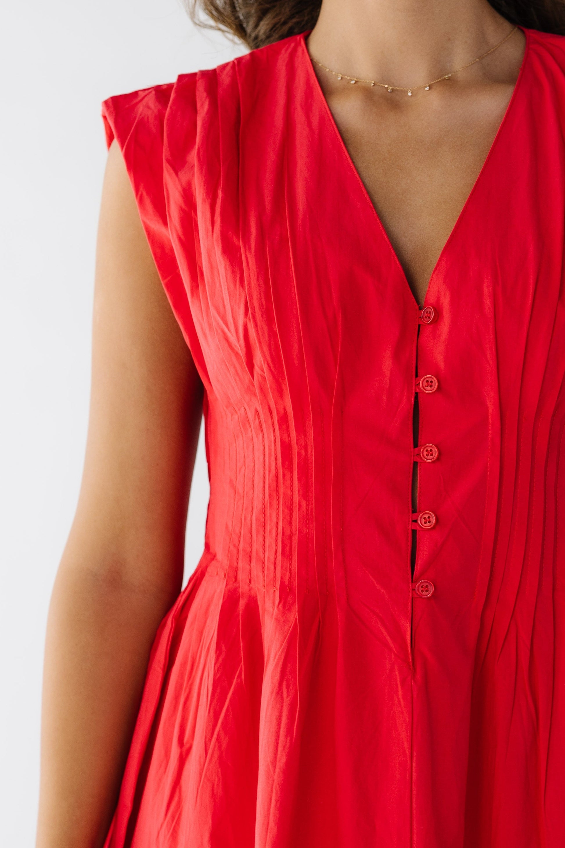 Closer view of a young woman wearing a red sleeveless dress with pleats at the shoulders and all around the waist with a short button placket with loop buttonholes.