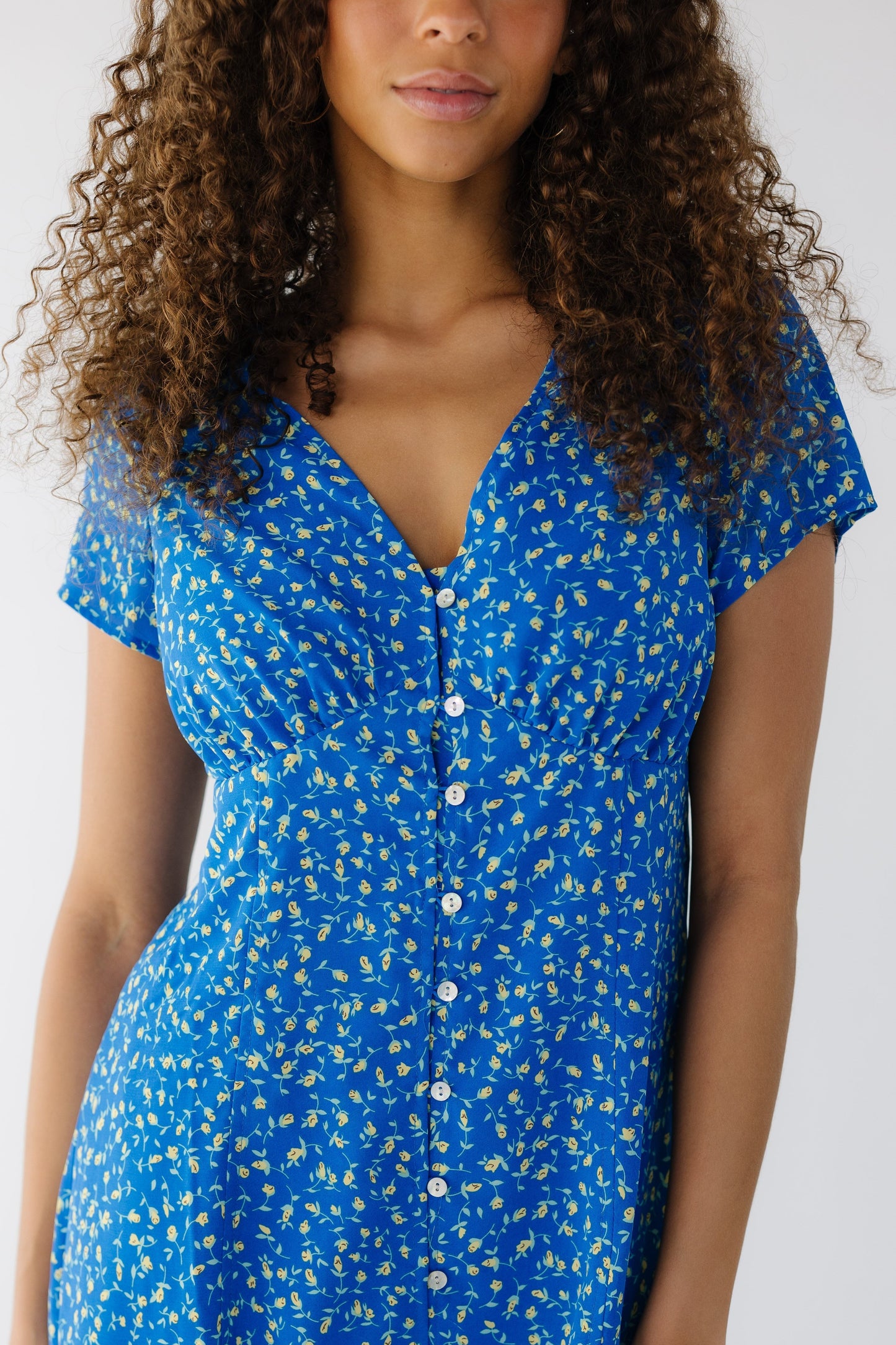 Close view of a a teen girl wearing a blue floral dress that buttons down the front with gathers under the bust with short sleeves and a V-neckline.