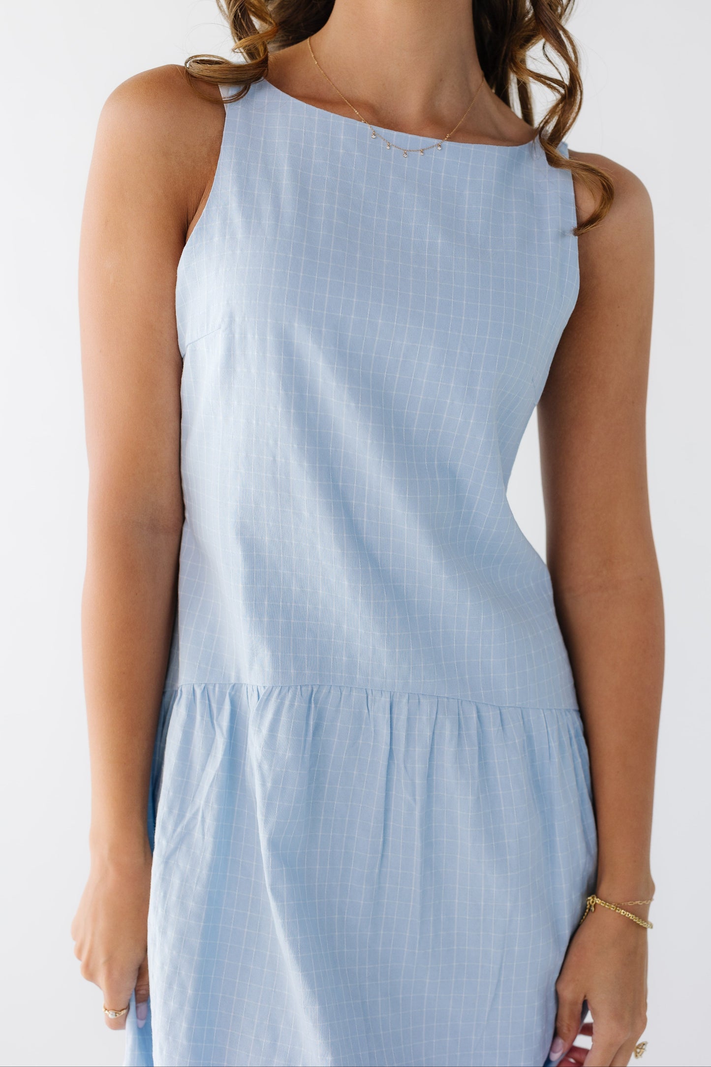 Close up view of a young woman wearing a light blue plaid sleeveless dress with a drop waist.