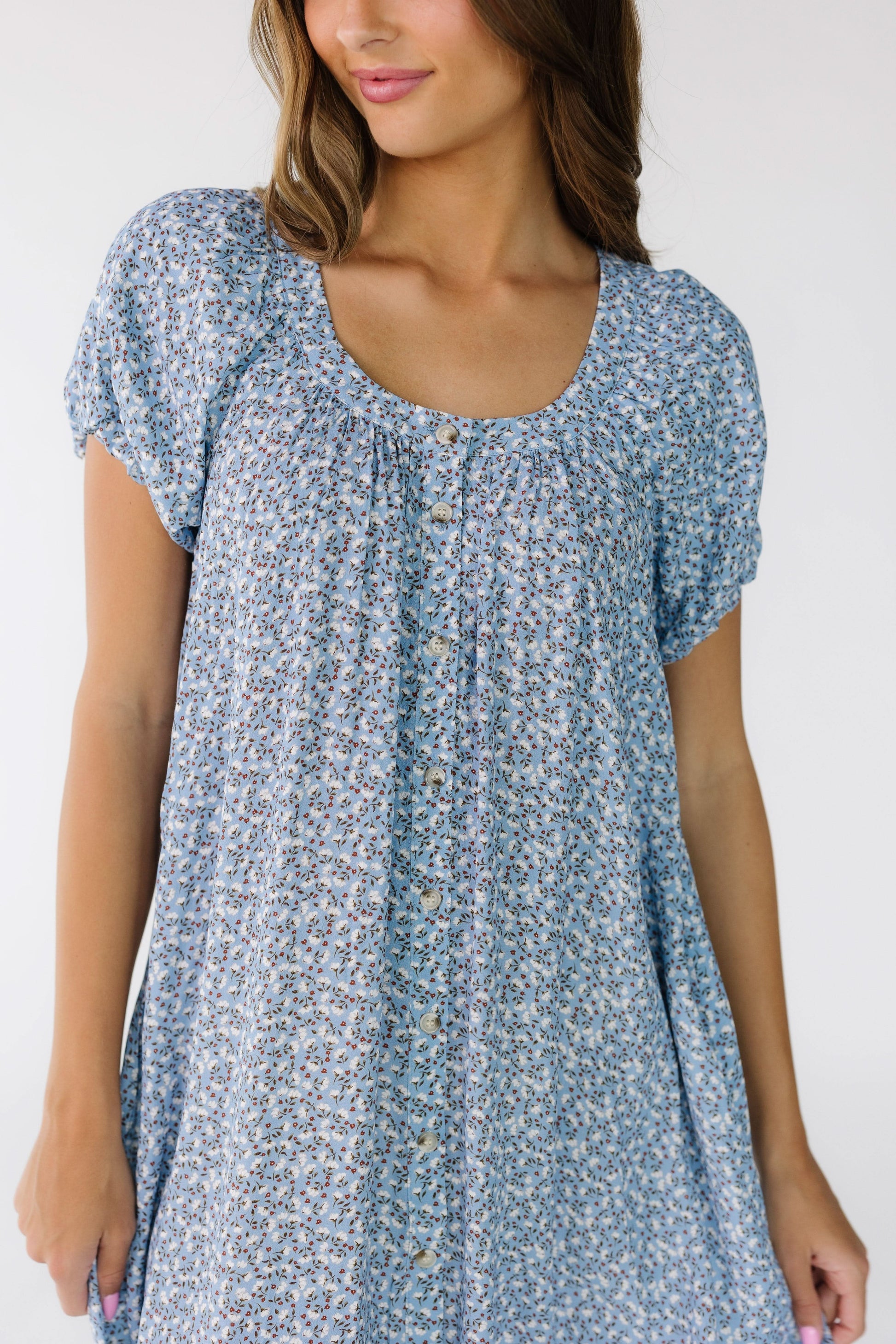 Close view of a young woman wearing a  blue floral button down dress with a banded scoop neckline and short puff sleeves.