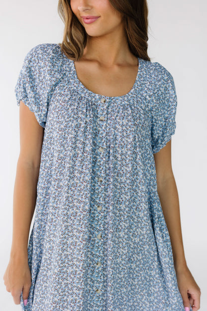 Close view of a young woman wearing a  blue floral button down dress with a banded scoop neckline and short puff sleeves.