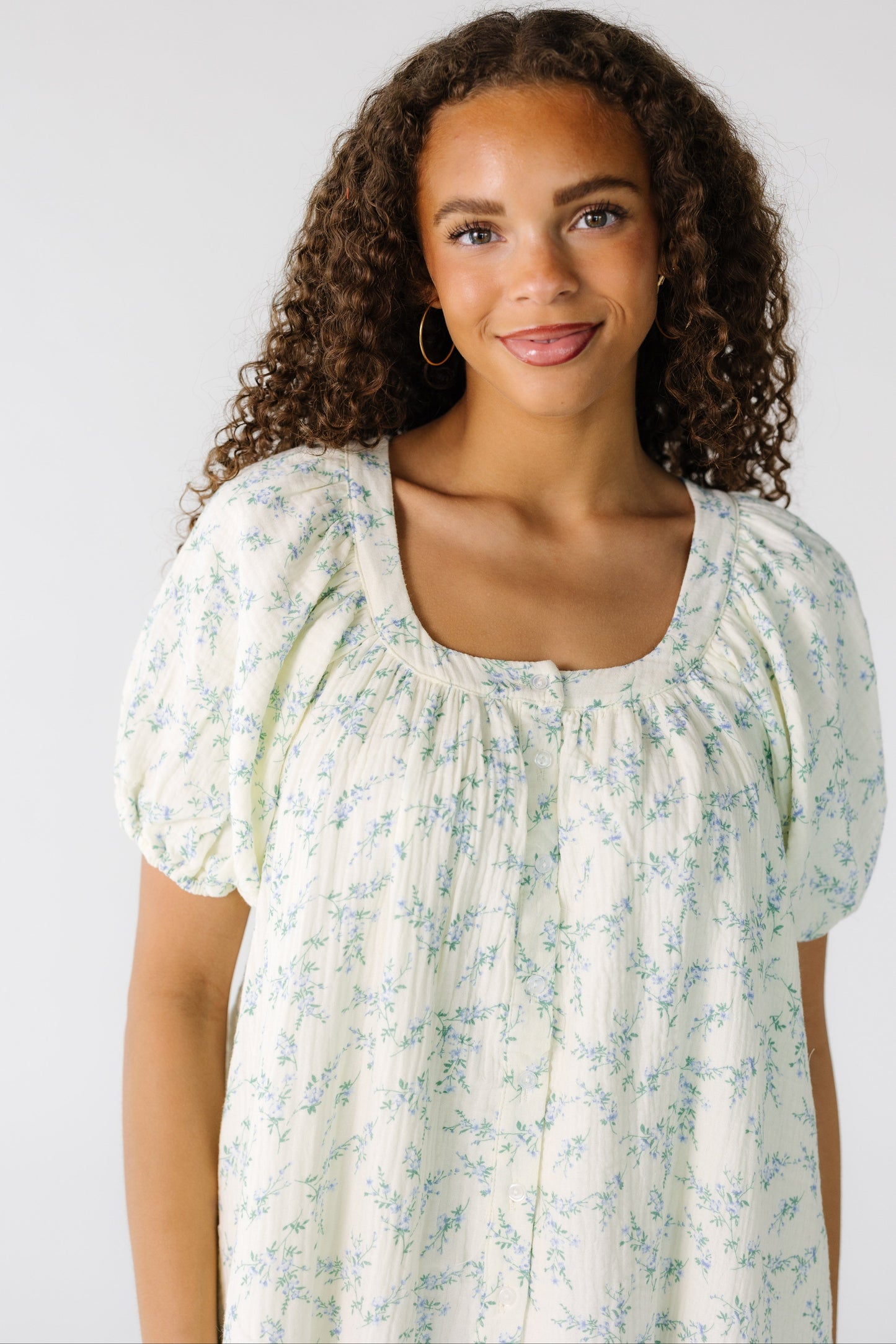 Close view of a teen girl wearing a cream loose fitting dress in a blue floral print with a scoop neckline and short puff sleeves.