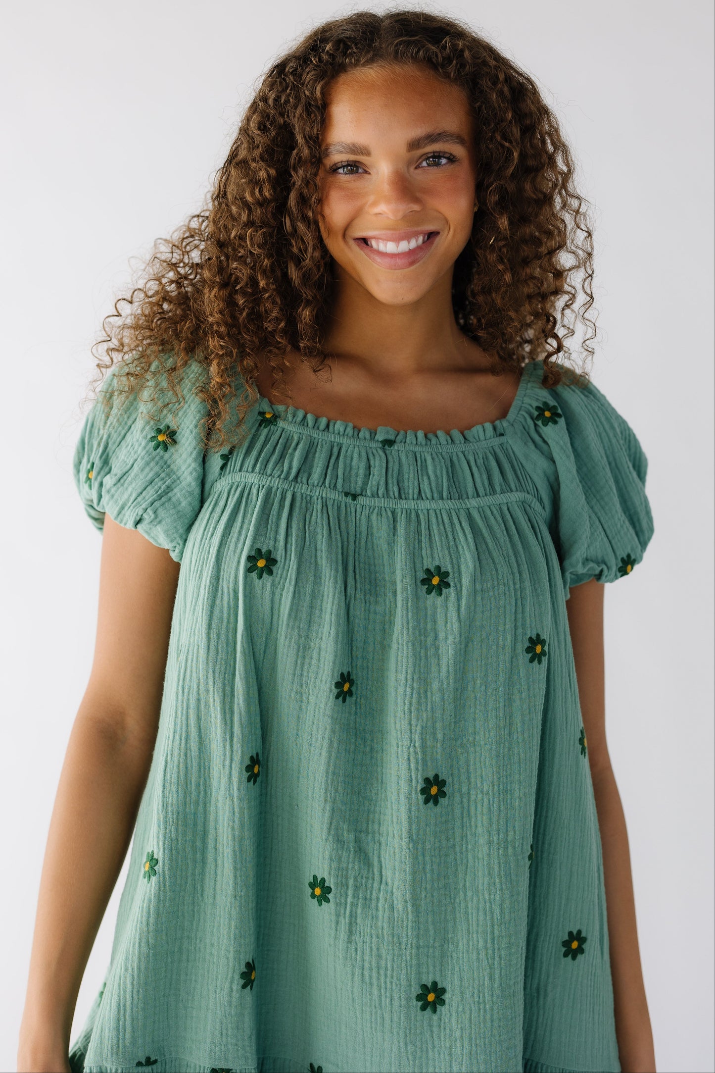 Close up view of a teenage girl wearing a loose fitting green dress with scattered green embroidered daisy's with large puff sleeves.