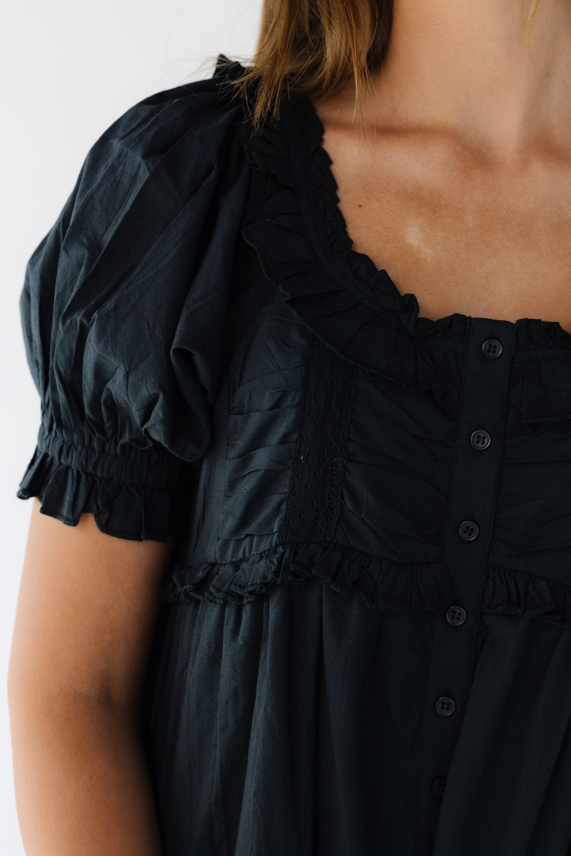 Close view of a woman wearing a black button down blouse with a scoop ruffled neckline, short puff sleeves.