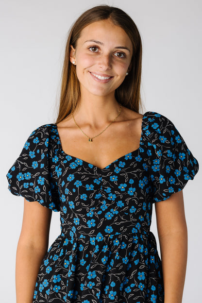 Close view of a woman wearing a black with blue floral dress with a sweetheart neckline and short puff sleeves