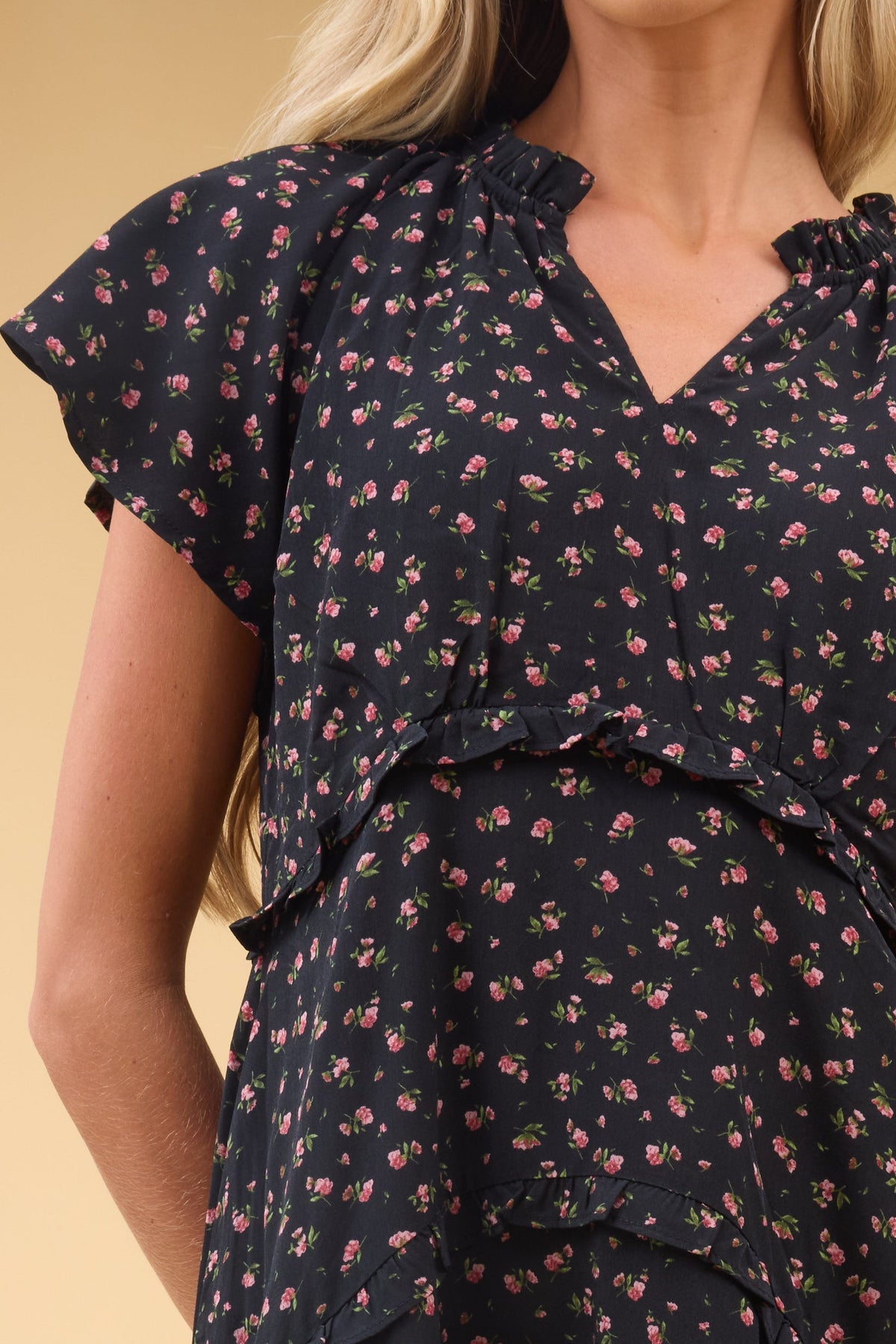 Close view of a woman wearing a black floral dress wtih a V-neckline, narrow ruffles and short flutter sleeves.