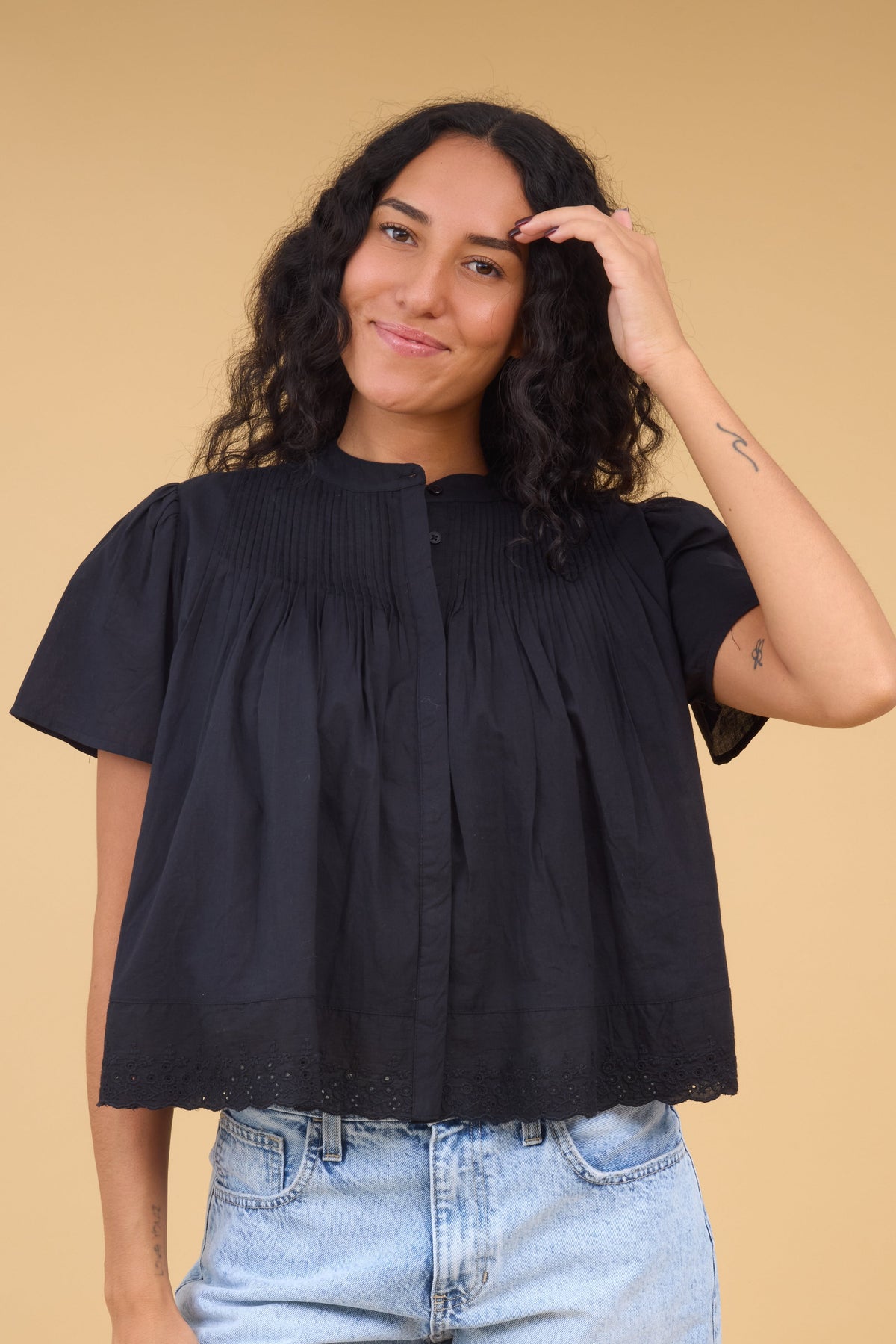 Young woman wearing a loose fitting black button down blouse with a hidden button placket, pintucks at the shoulders and short sleeves.