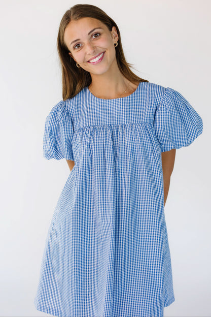Young woman wearing a blue and white gingham short dress with large puff sleeves.