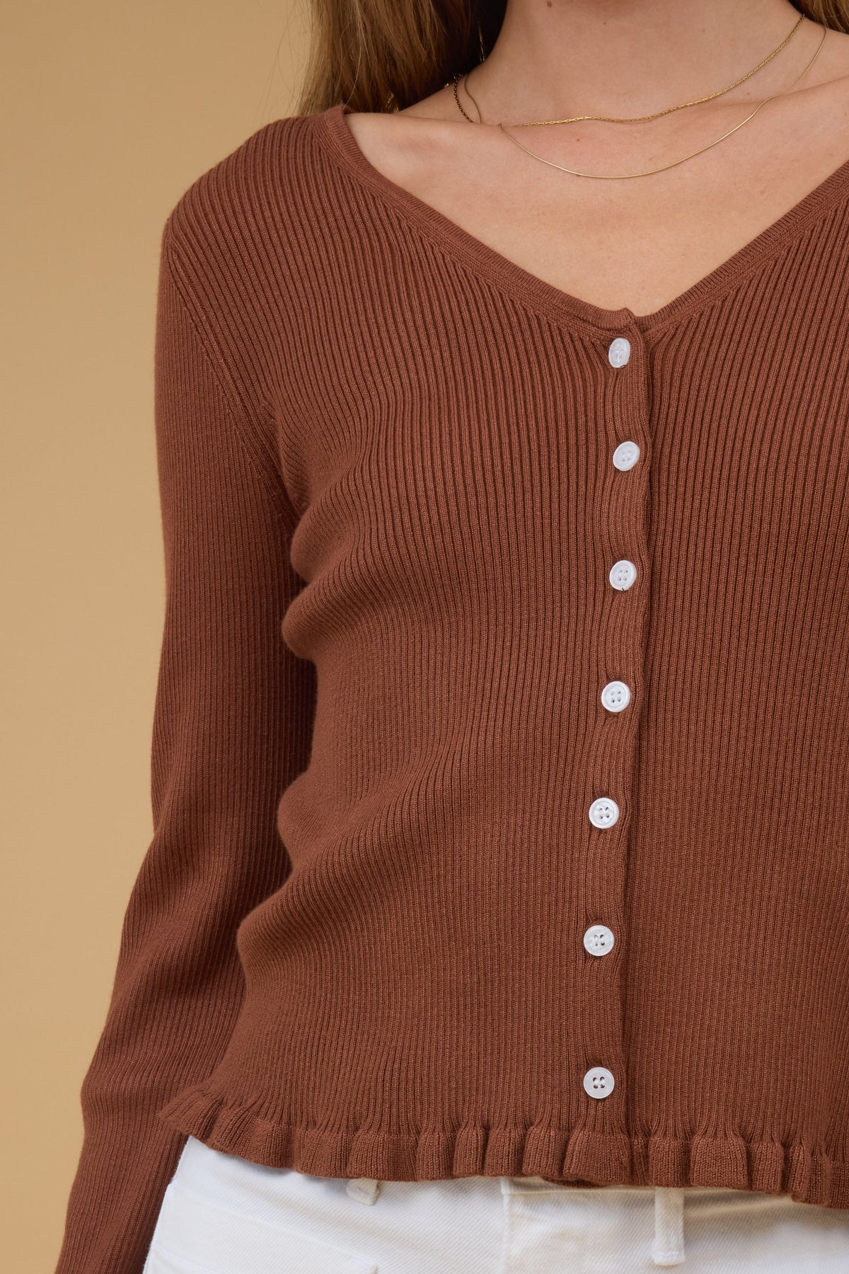 Close view of a woman wearing a rib knit button down top in brown with a V-neckline, contrastingbuttonsand a ruffle hem.