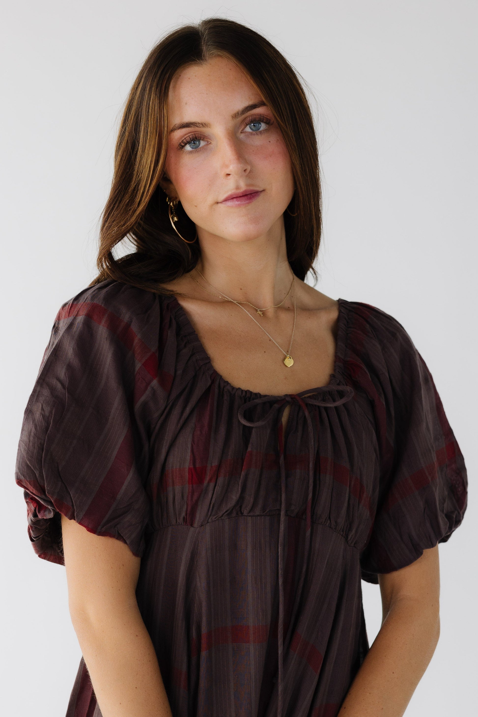 Close bodice view of a young woman wearing a brown and red plaid dress with a drawstring-scoop neckline, empire waist and ballong sleeves.