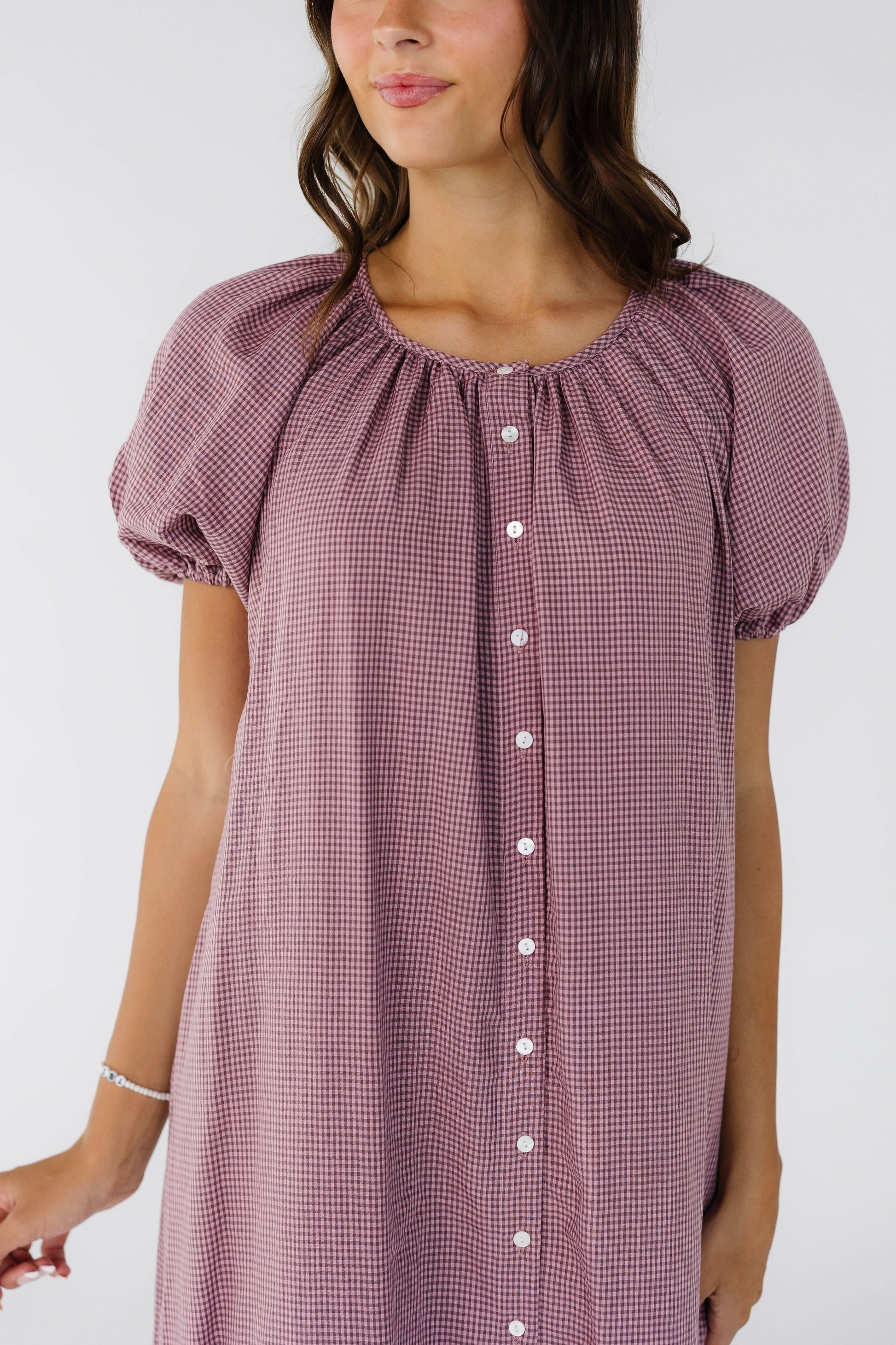 Close view of a woman wearing a button down dress in burgundy & pink gingham with a slight scoop neckline and puff sleeves.