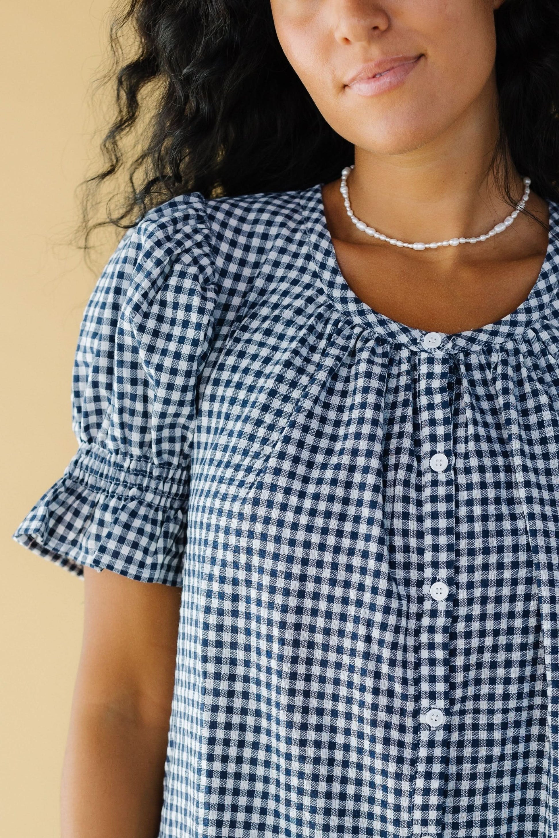 Close view of a woman wearing a navy & white gingham button down dress with a scoop neckline and puff sleeves with elastic & ruffled cuffs.