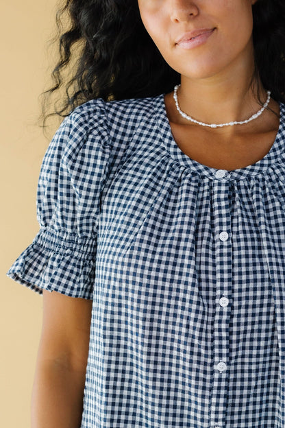 Close view of a woman wearing a navy & white gingham button down dress with a scoop neckline and puff sleeves with elastic & ruffled cuffs.