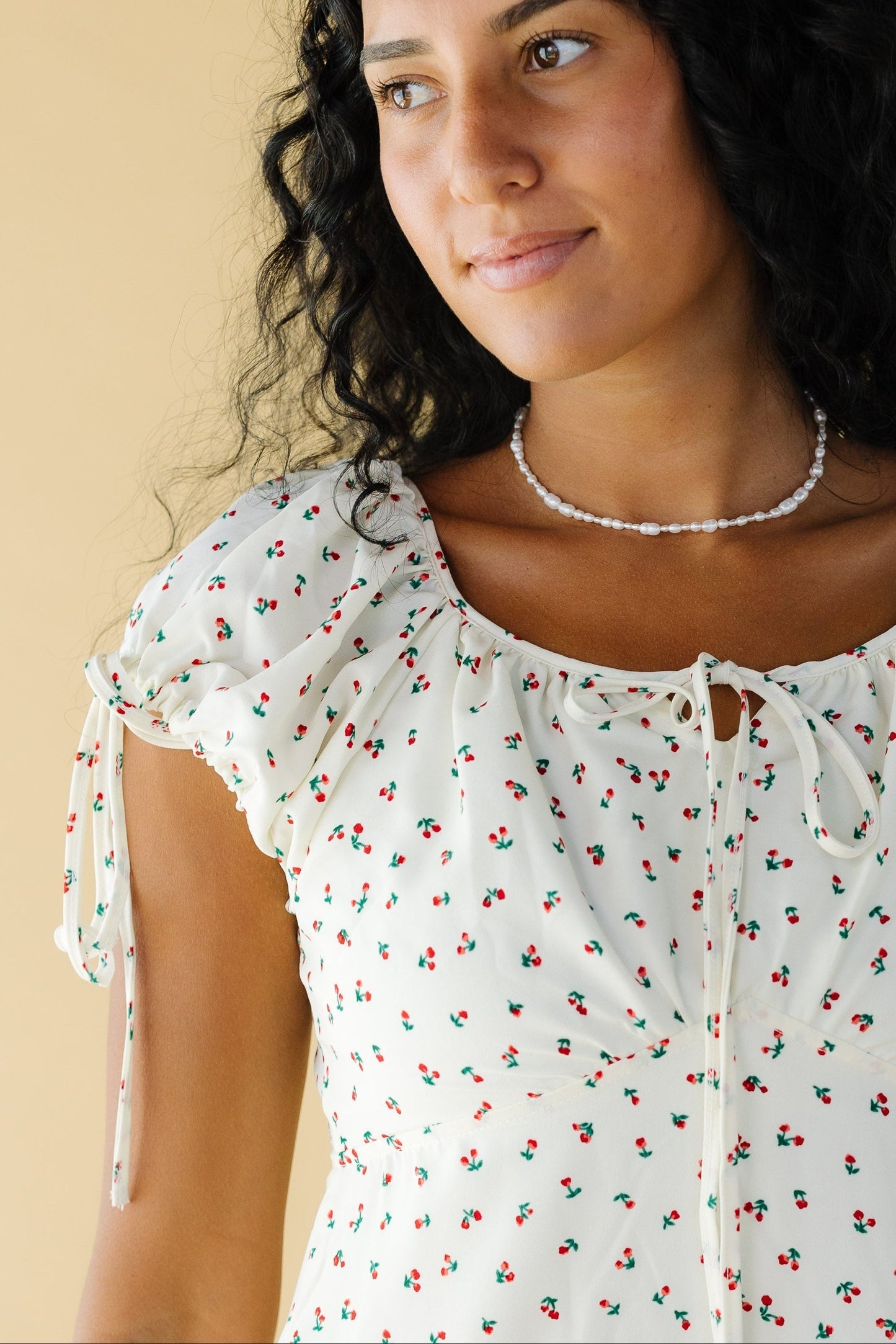 Close up view of a woman wearing a cream dress with red floral print, a wide round neckline with a bow and short puff sleeves including a tie cuff.