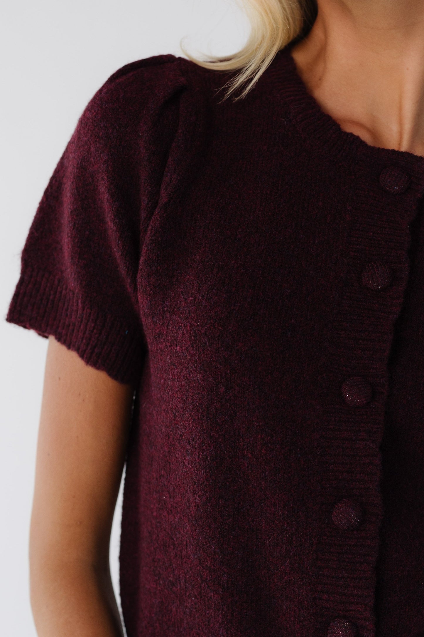 Close up view of a woman wearing a burgundy button down cardigan with matching covered buttons, scallop hems & neckline and short sleeves.