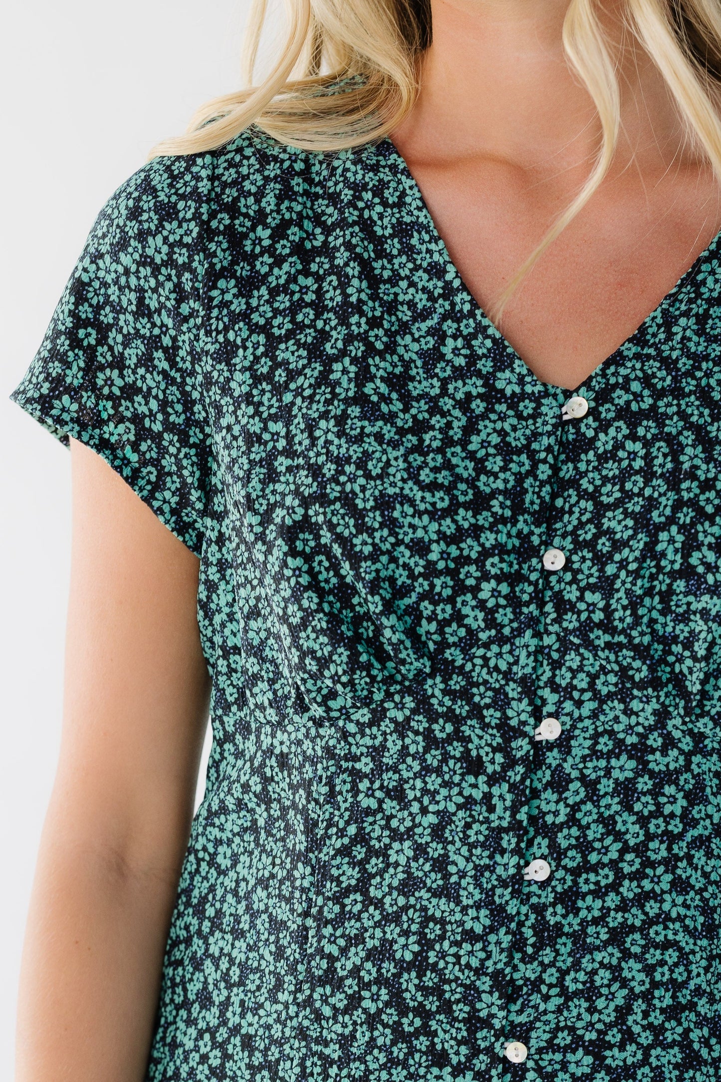 Close view of a woman wearing a button down green floral dress with a V-neckline, short sleeves and an empire waist.