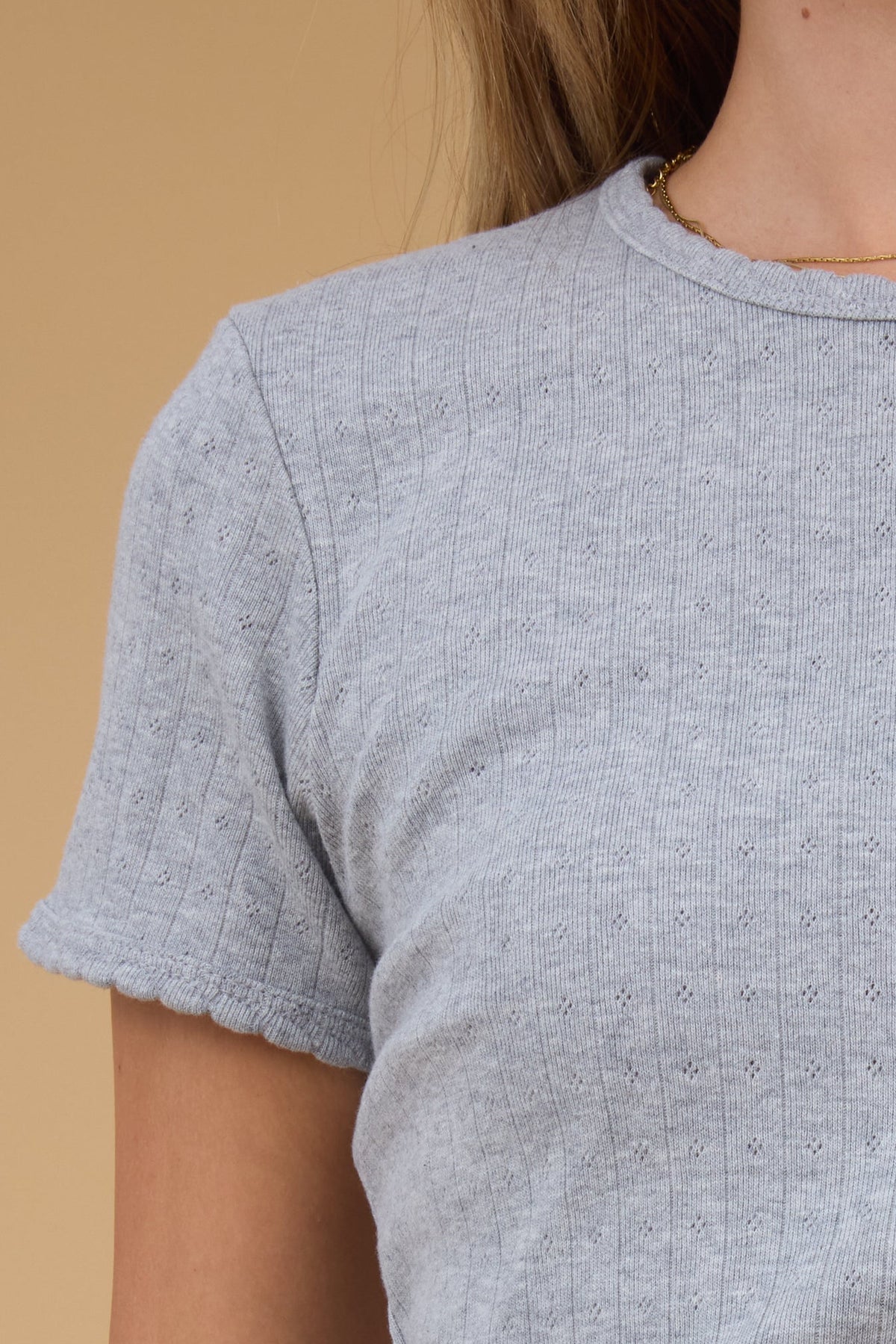 Close view of a woman wearing a grey, close fitting pointelle knit top with a scallop edge round neckline and short sleeves with a scallop hem. 