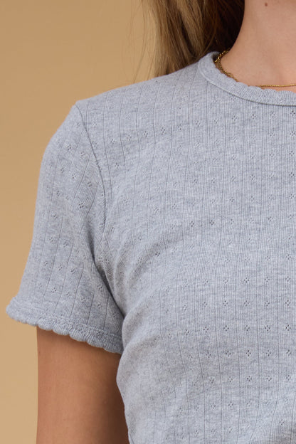 Close view of a woman wearing a grey, close fitting pointelle knit top with a scallop edge round neckline and short sleeves with a scallop hem. 