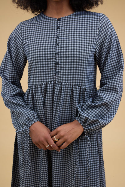 Close view of a woman wearing a blue gingham dress with a round neckline, long sleeves with ties and a button down bodice with elastic loop button holes.