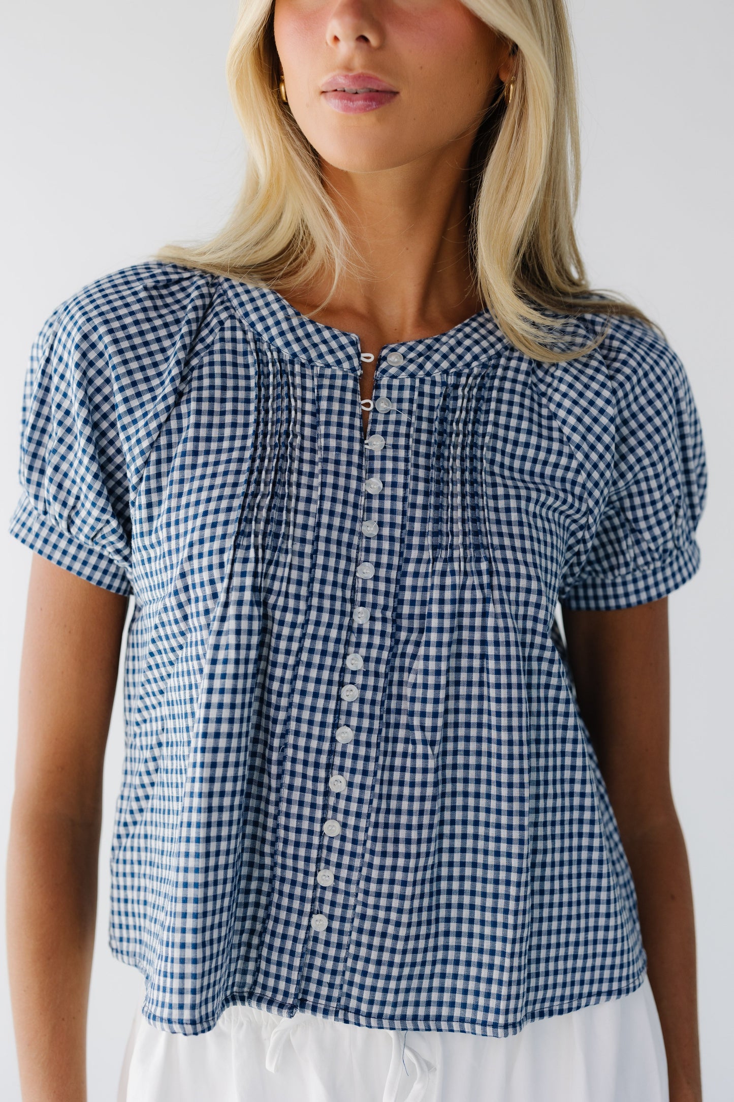 Close view of a young woman wearing a navy & white button down gingham blouse with a round neckline and short puff sleeves.
