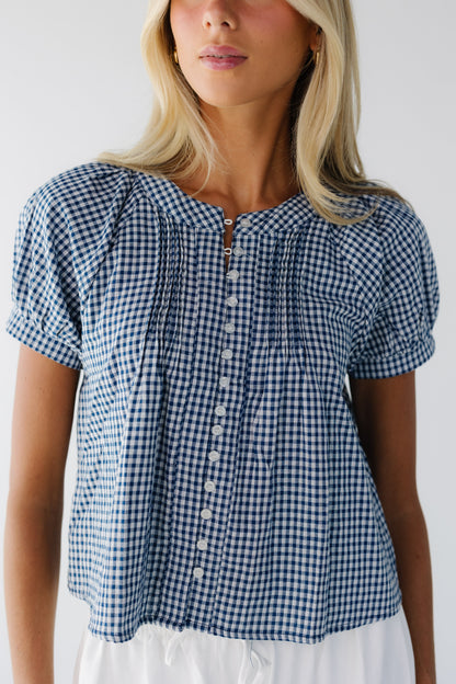 Close view of a young woman wearing a navy & white button down gingham blouse with a round neckline and short puff sleeves.