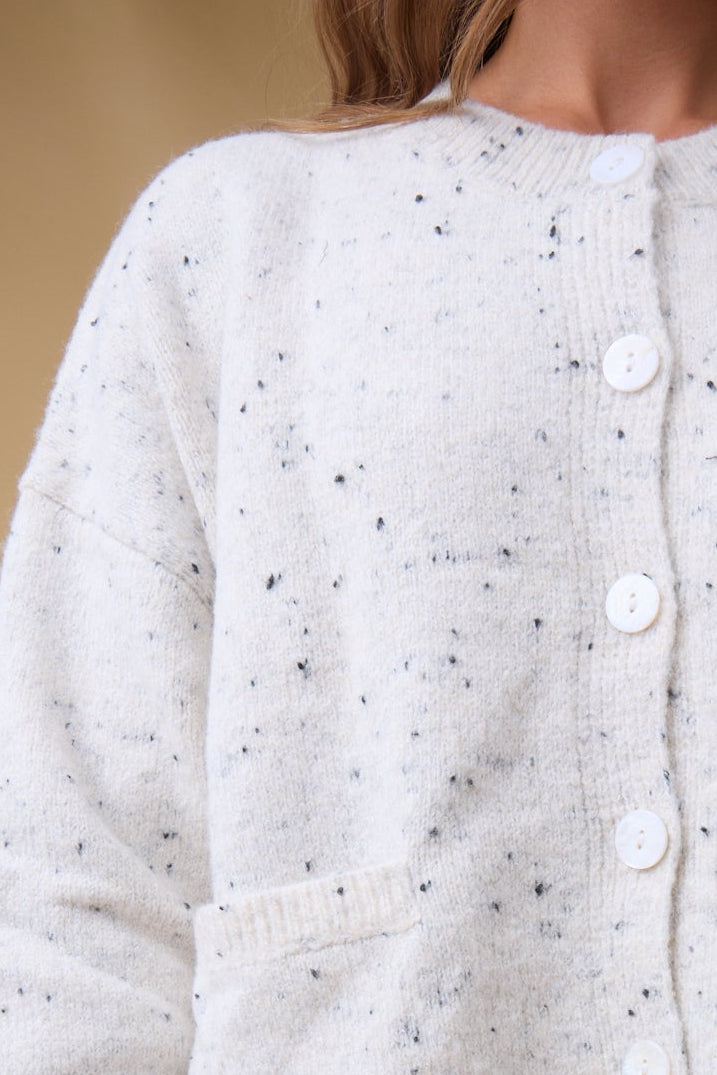 Close view of a young woman wearing a cream flecked, button down cardigan with a crew neckline, long sleeves and front pockets.