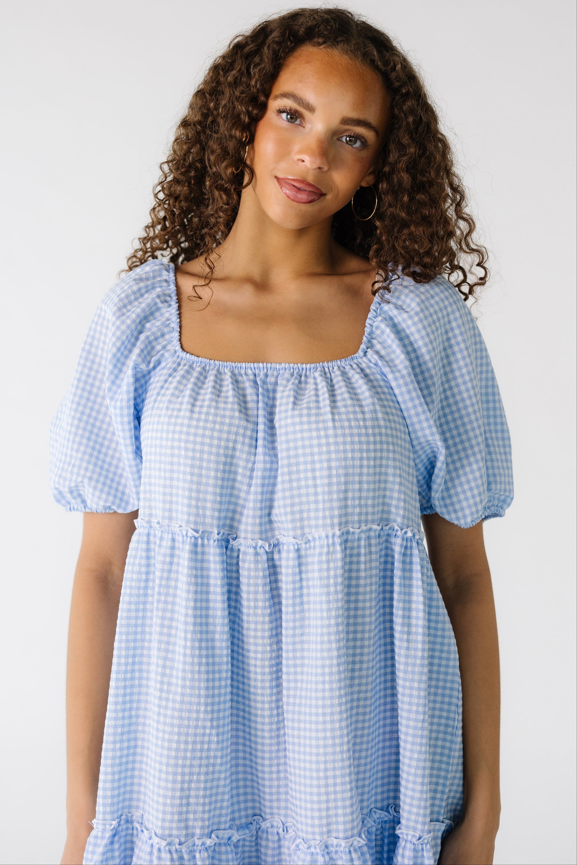 Close view of a teen girl wearing a light blue gingham dress with a square neckline and short puff sleeves.