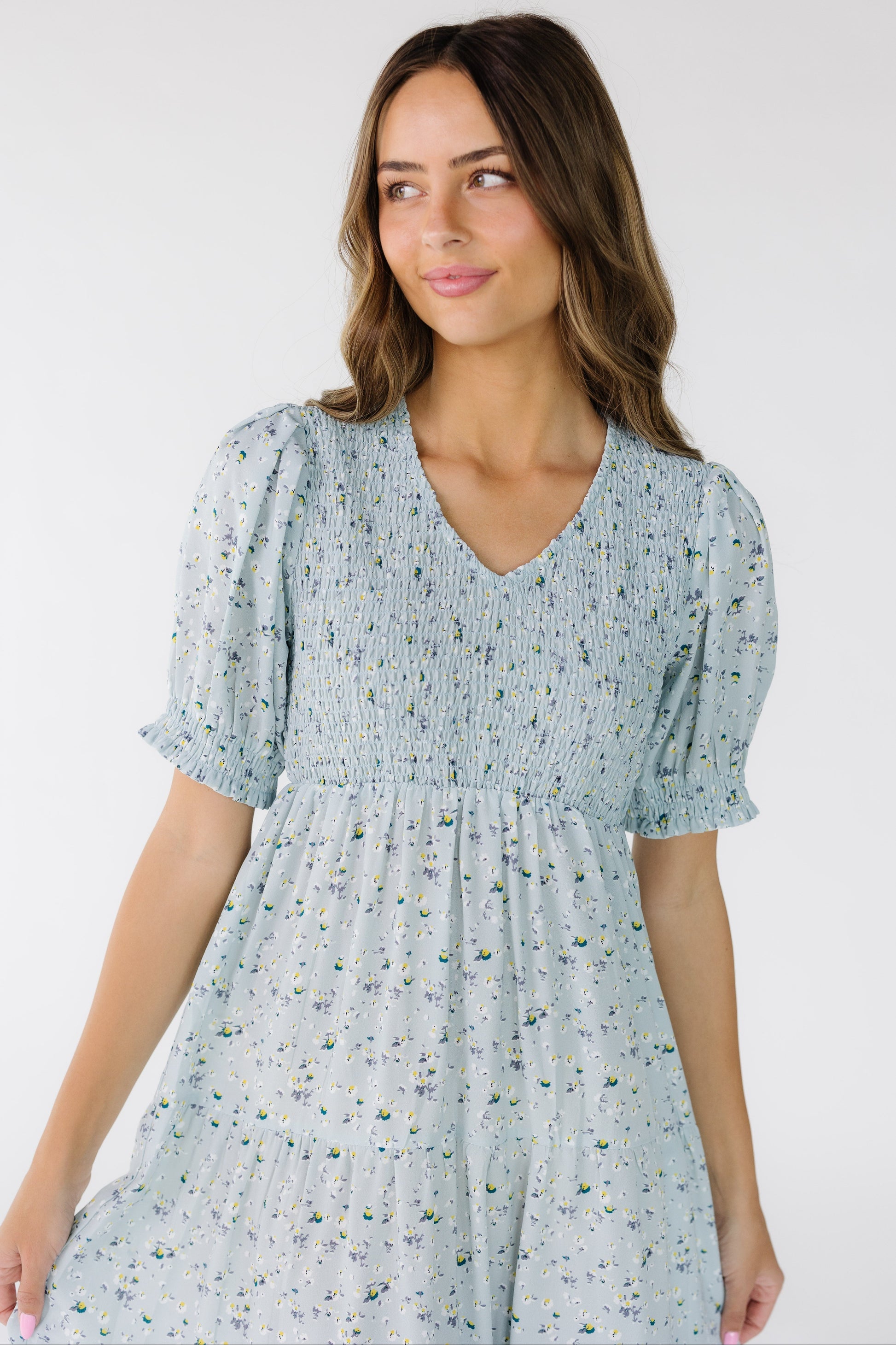 Close view of a  young woman wearing a light blue floral midi with a front and back smocked bodice with a V-neckline with short sleeves with a ruffle hem.