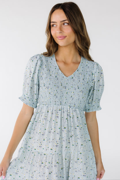 Close view of a  young woman wearing a light blue floral midi with a front and back smocked bodice with a V-neckline with short sleeves with a ruffle hem.